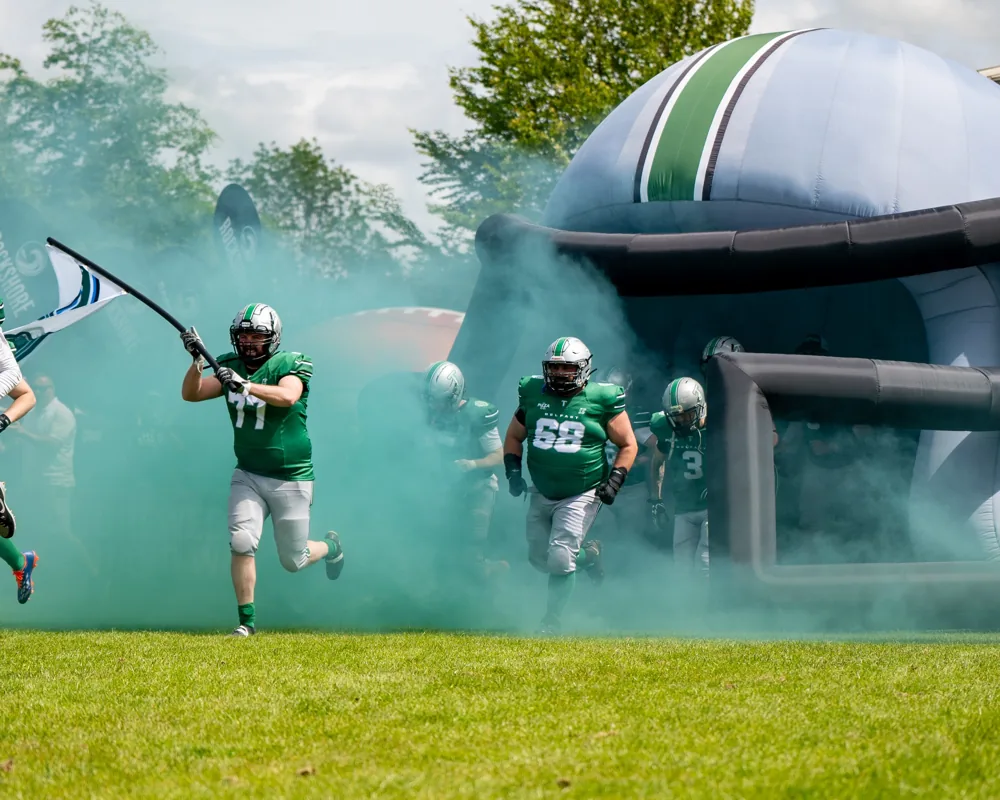 Belfast Trojans players run through green smoke beside an inflatable helmet.